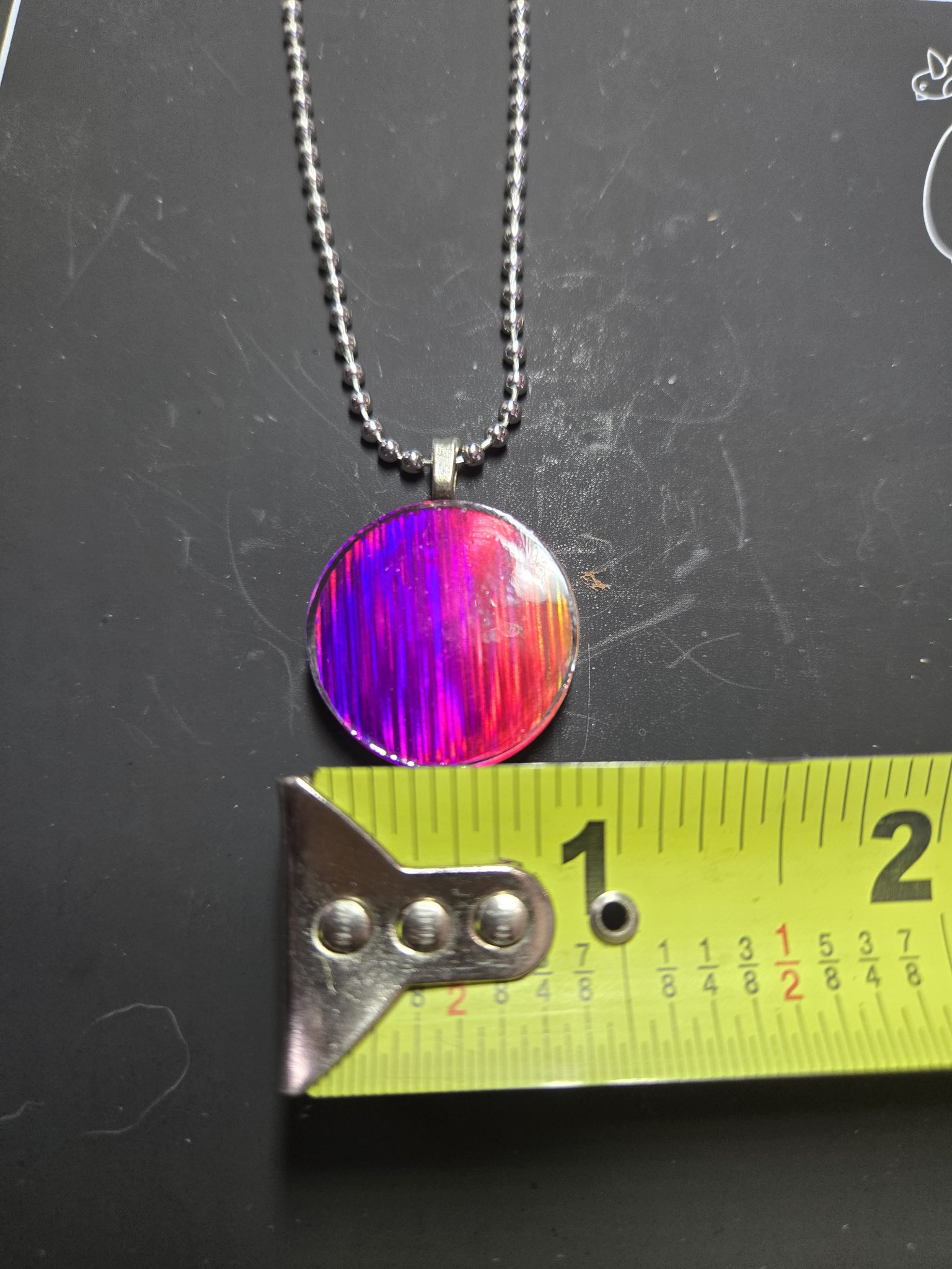 Magenta side of the holographic pendant against a tape measure showing it is about one and a half inches across
