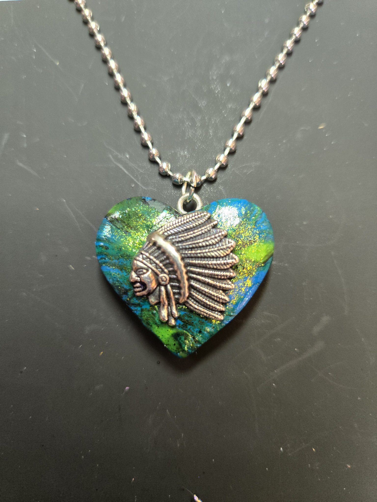 Front of a green, blue, and gold heart-shaped polymer clay pendant with a silver-tone Native American headdress chief charm on a ball chain
