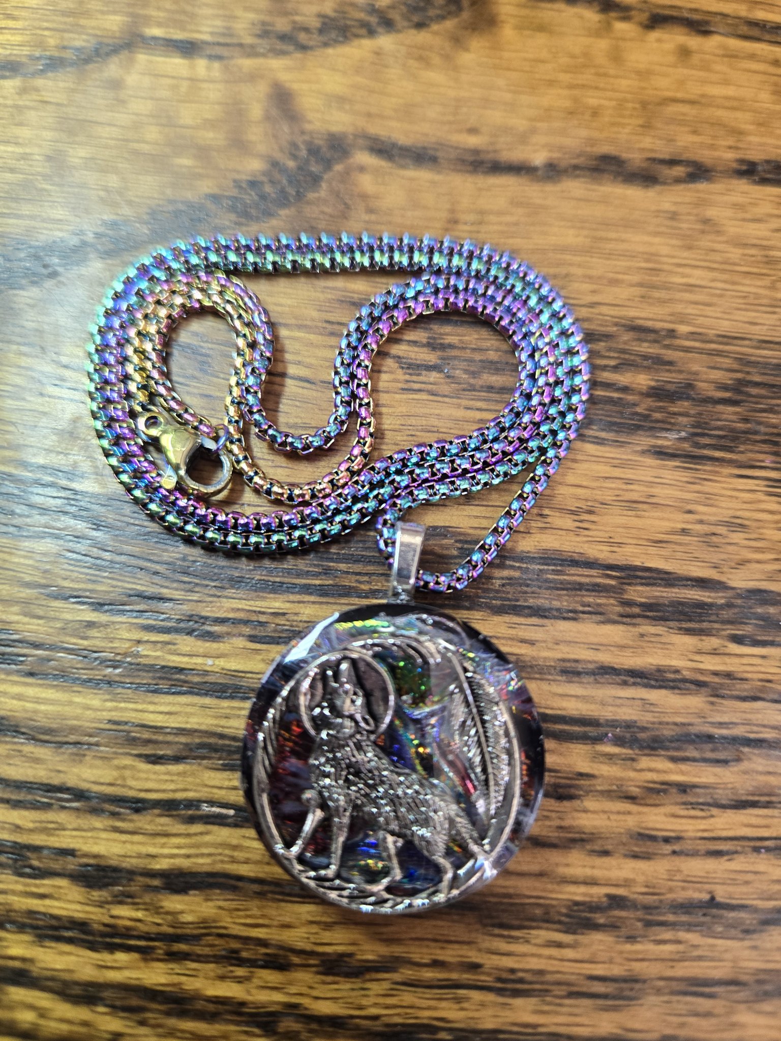 Front of a round holographic resin pendant with a silver-tone howling wolf, moon, and feather design on a rainbow iridescent chain