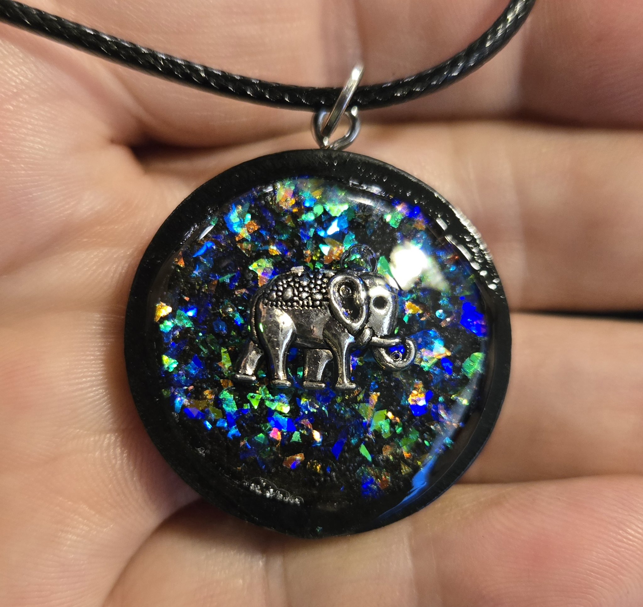 Close-up of a round holographic resin pendant with blue, green, and gold flakes, a silver-tone elephant charm, and black frame, held in a hand
