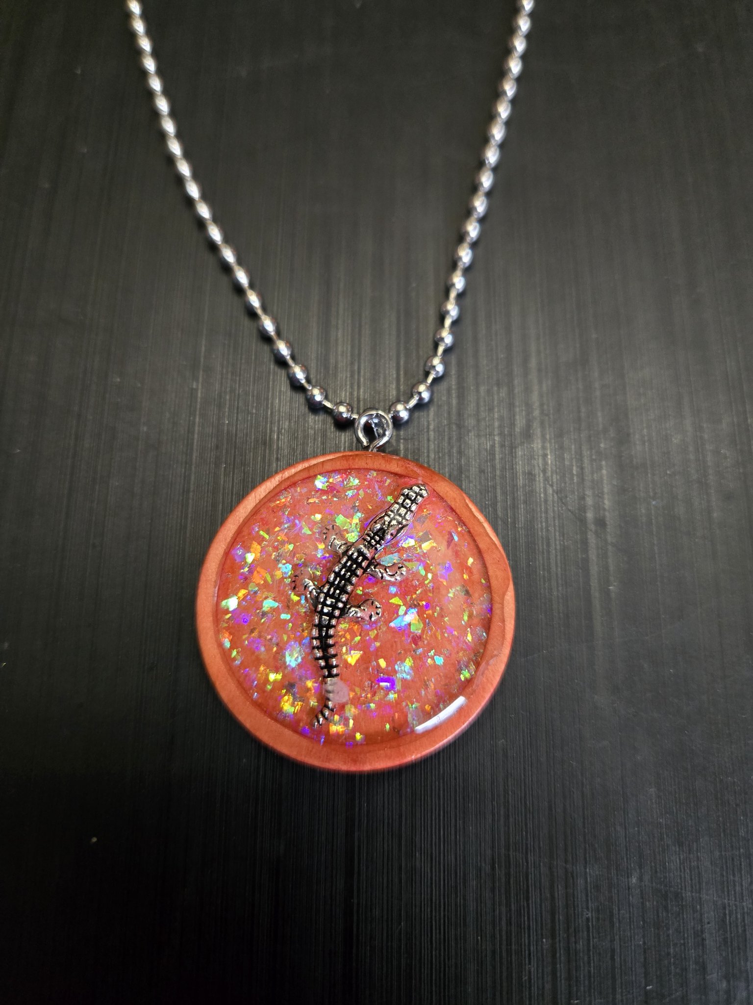 Front view of a round orange-pink wood pendant with rainbow holographic glitter, a silver-tone alligator charm, and a ball chain