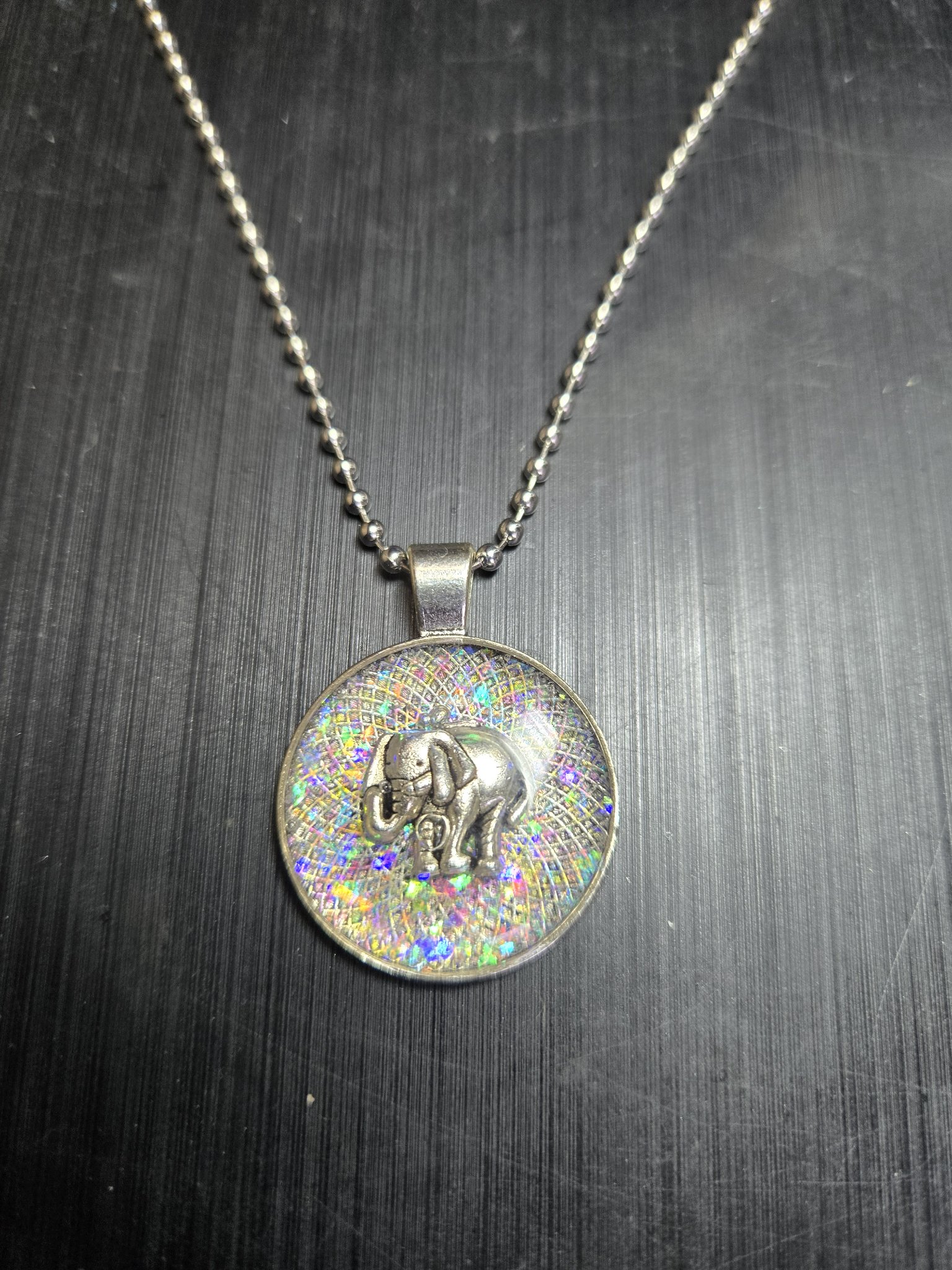 Front view of the round metal and resin pendant with holographic glitter inlay, a silver-tone elephant charm, and a ball chain