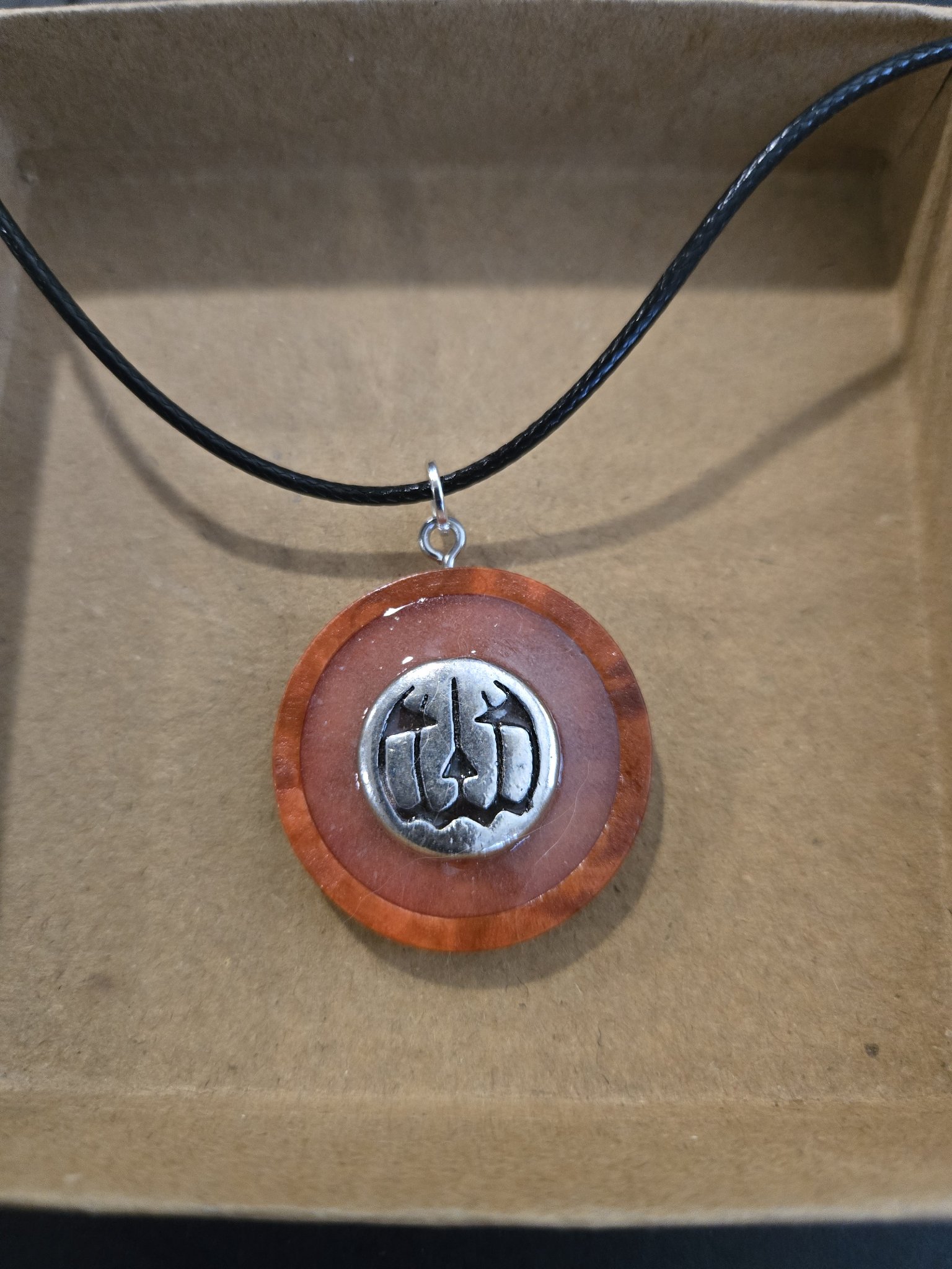 Stock 166 round orange and pink resin pendant with wood backing, a silver-tone jack-o-lantern charm, and a black leather cord necklace in daylight