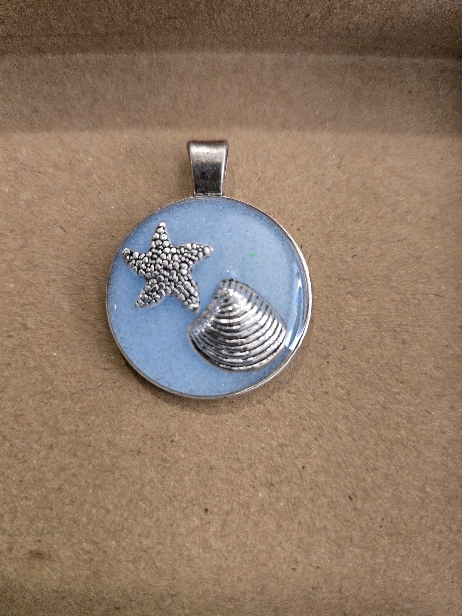 Stock 170 round light blue resin beach theme pendant in a silver metal frame with silver-tone starfish and seashell charms in daylight