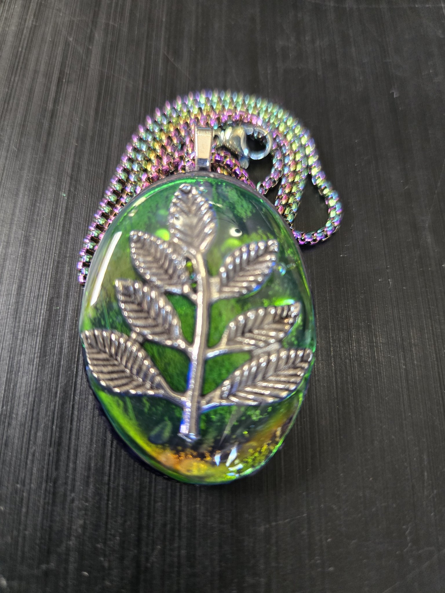 Front close-up of stock 193 oval green, blue, and gold iridescent holographic resin pendant with a silver-tone branch and leaf charm