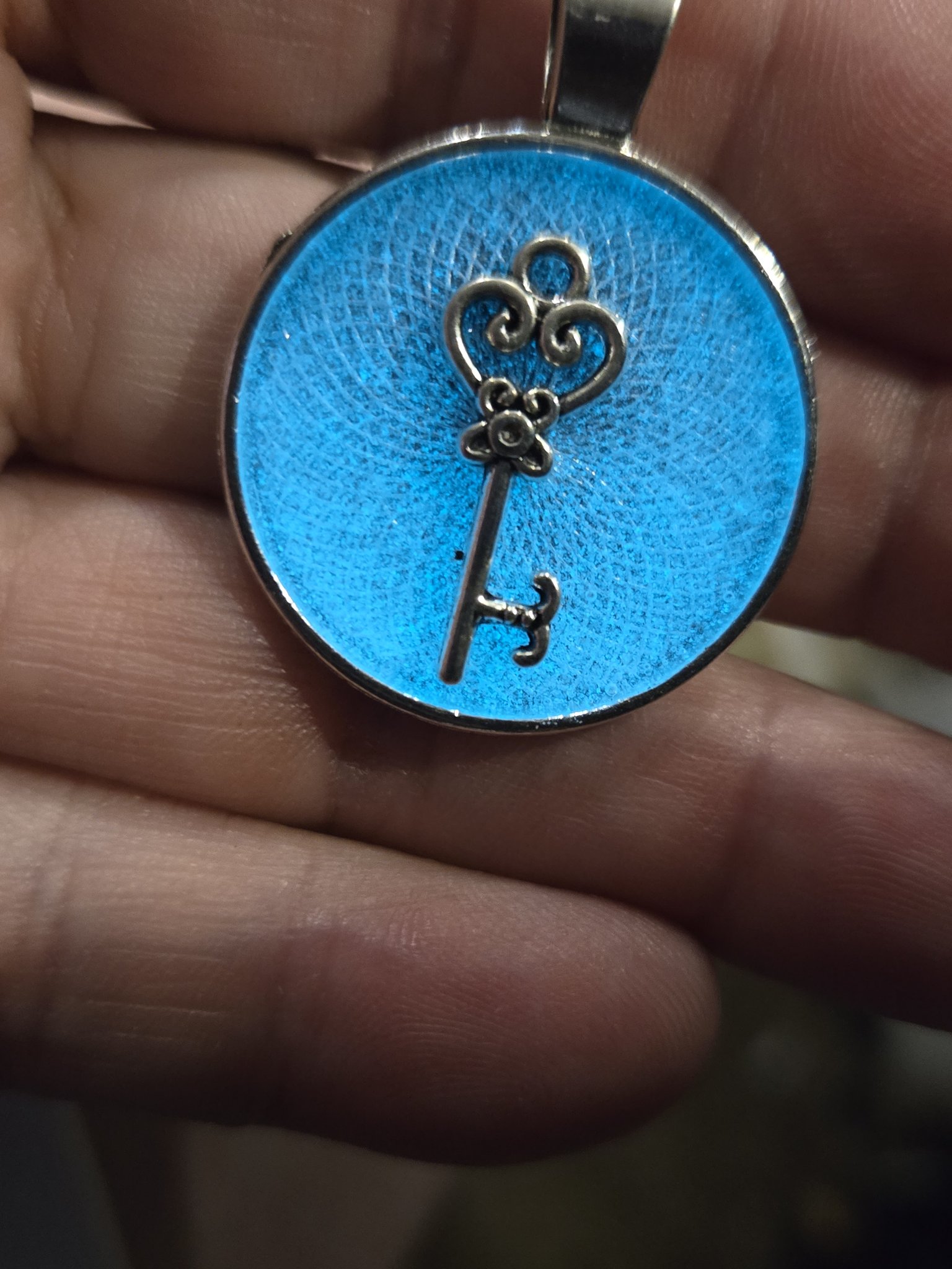 Stock 202 round bright blue glow-in-the-dark key resin pendant in a silver metal frame, shown from the front in a hand
