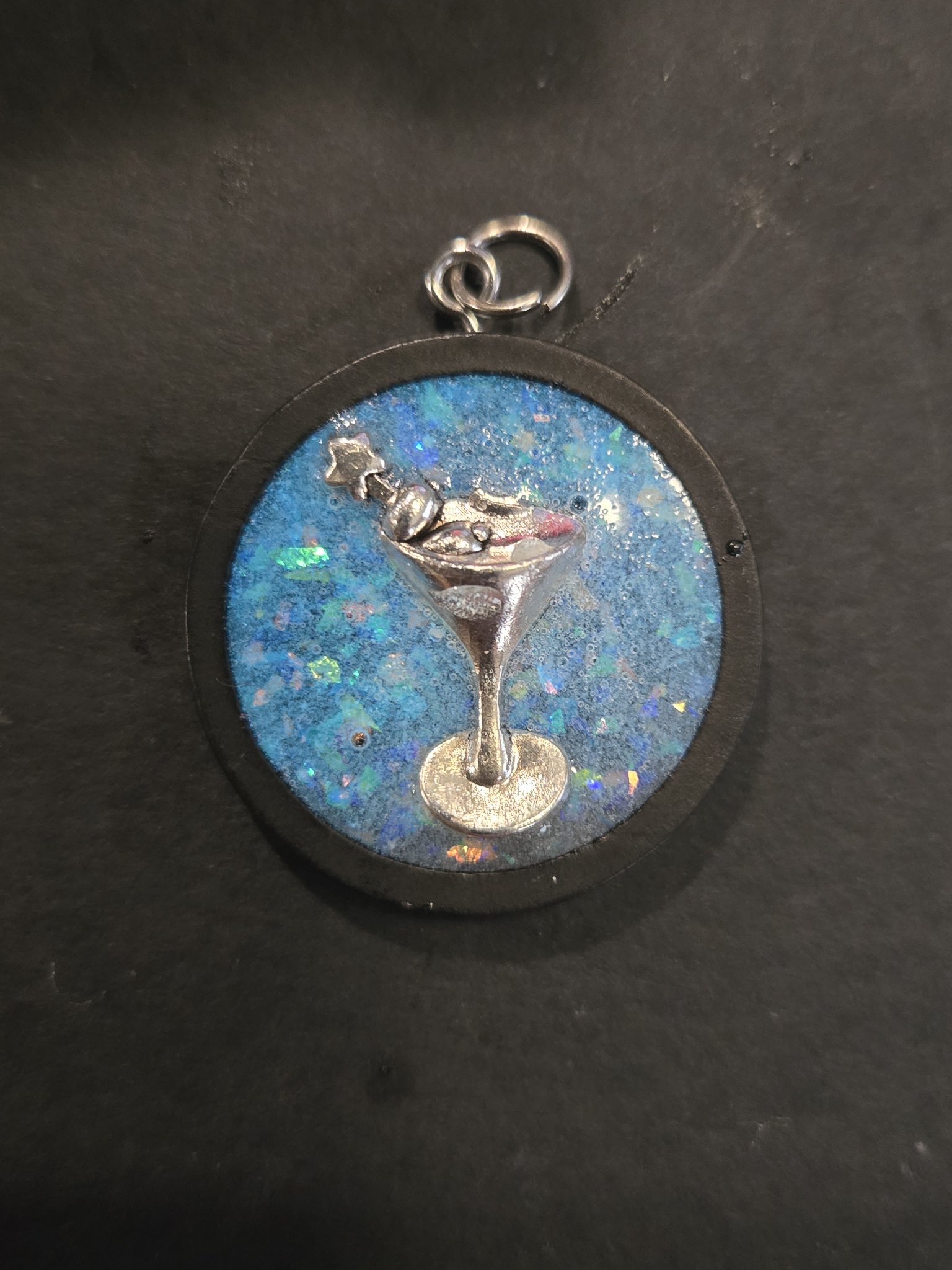 Stock 203 round blue glow-in-the-dark martini resin pendant in a black wood frame, shown from the front in a hand