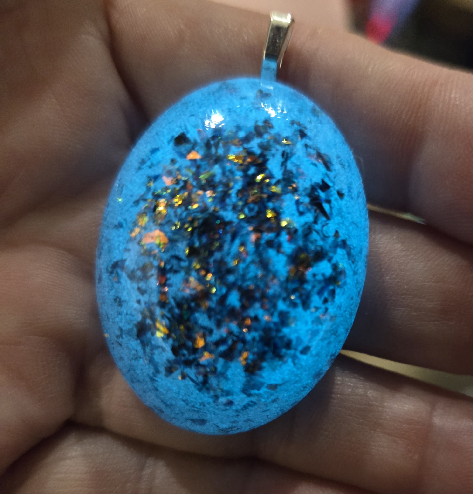 Stock 251 holographic glow oval pendant held in a hand, glowing a bright vivid blue with the holographic flecks showing through