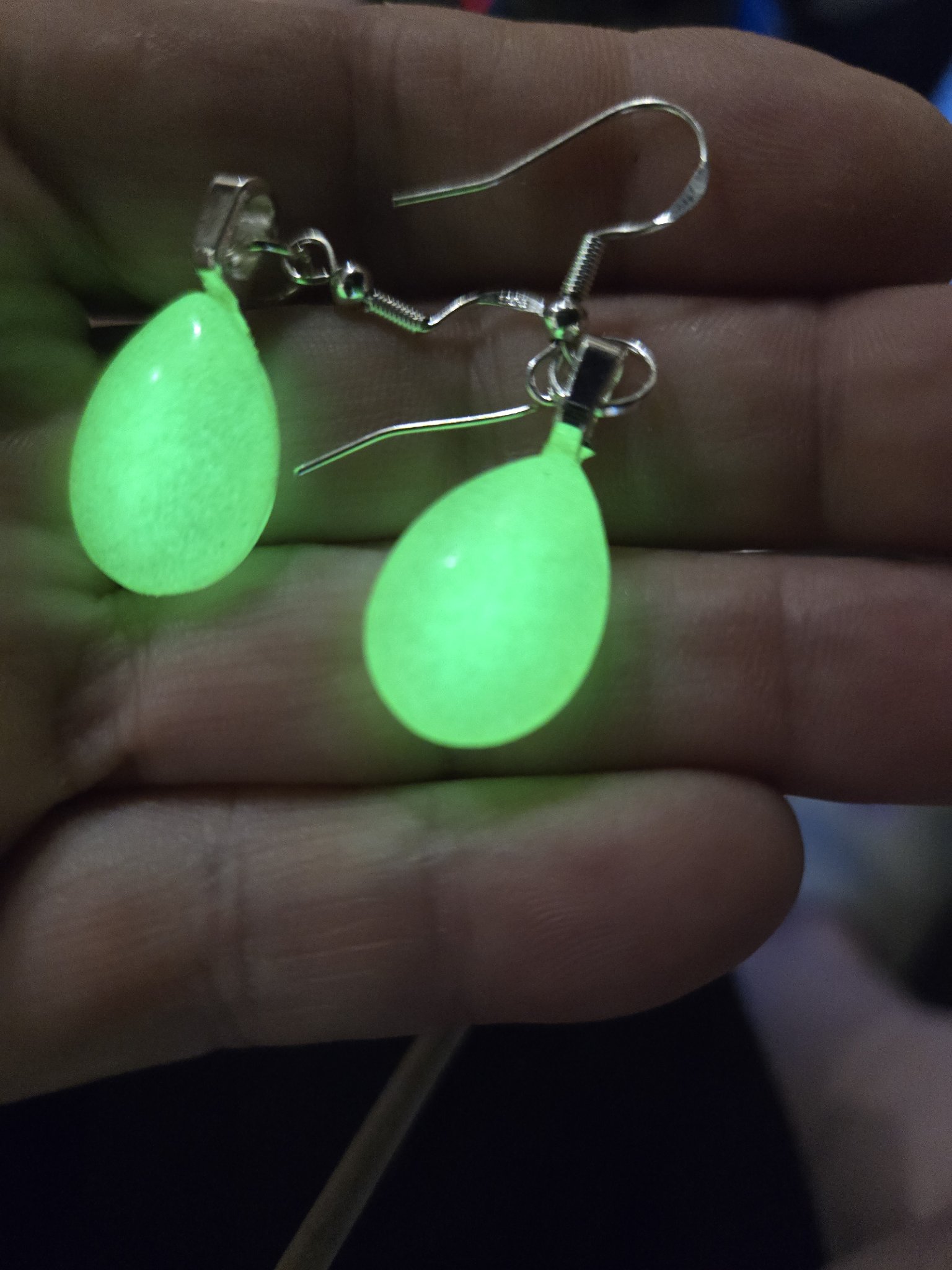 Stock 252 glow in the dark teardrop earrings held in a hand, the resin teardrops glowing bright green on sterling silver wires
