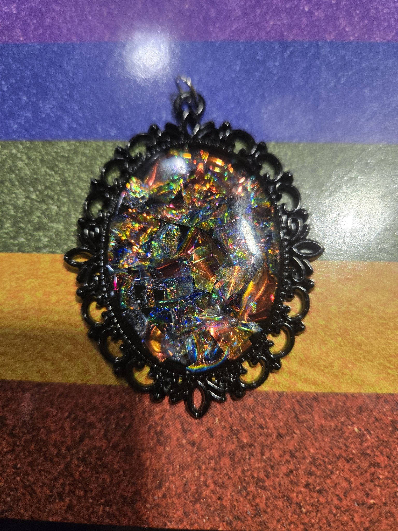 Stock 253 oval holographic filigree pendant shown from the front, rainbow holographic flakes under a domed resin cabochon in an ornate black metal filigree frame