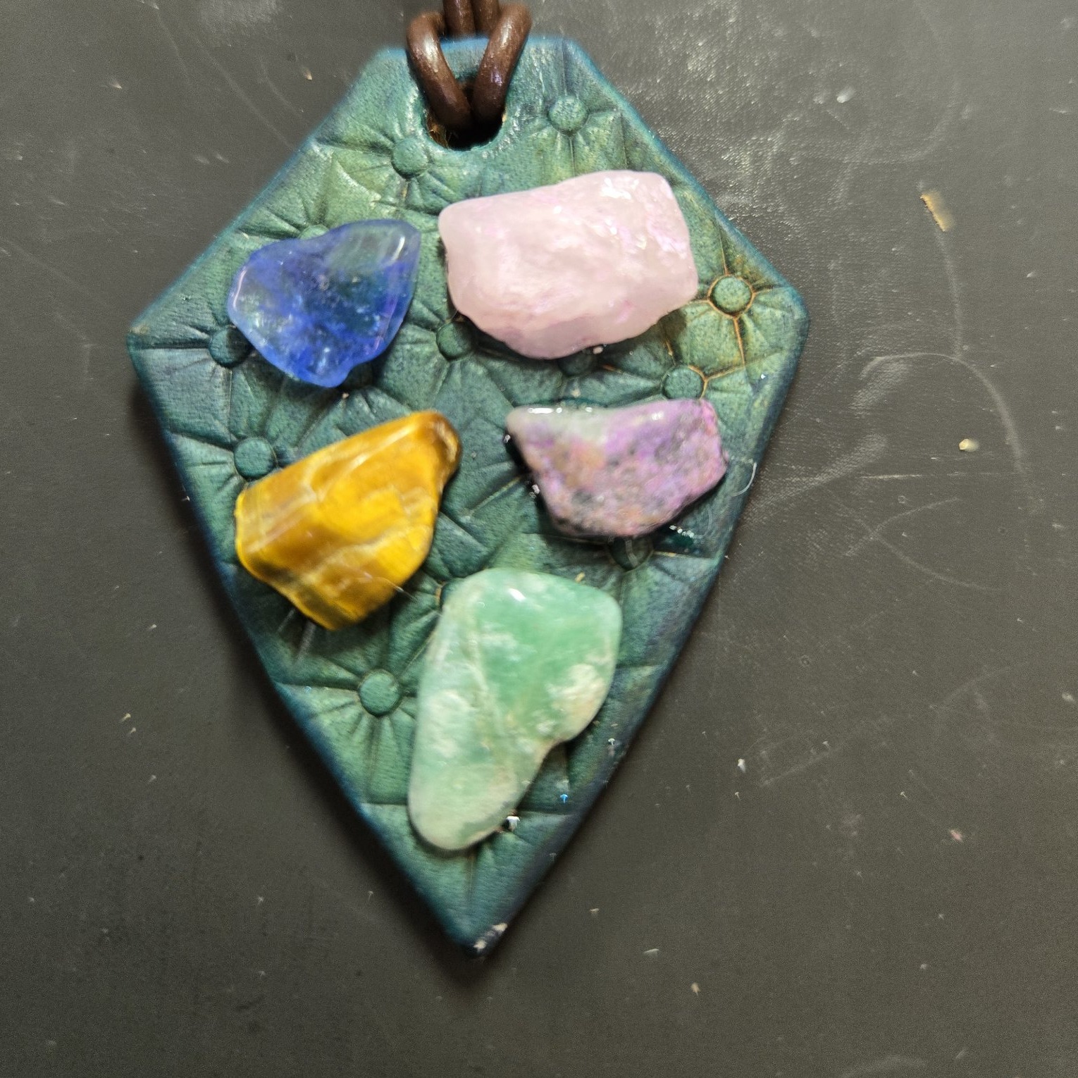 Close-up of a diamond-shaped teal vegetable-tanned leather pendant set with rose quartz, blue glass, tiger's eye, a purple stone, and green jasper