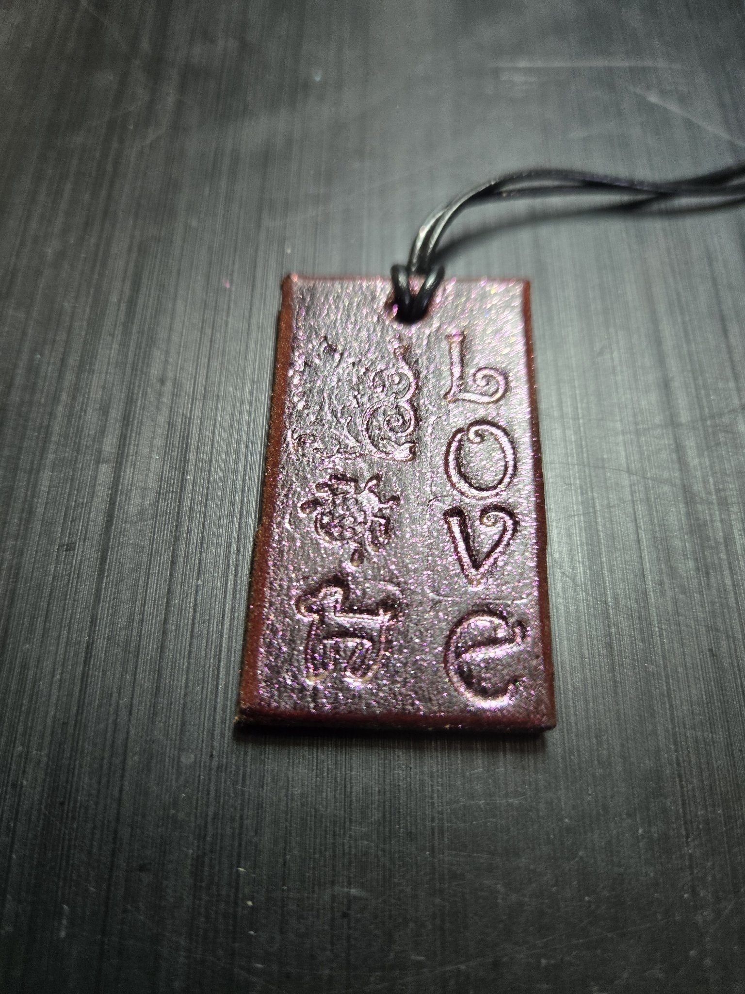 Front close-up of a rectangular pink sparkle leather pendant hand-stamped with LOVE and floral designs on a black leather cord