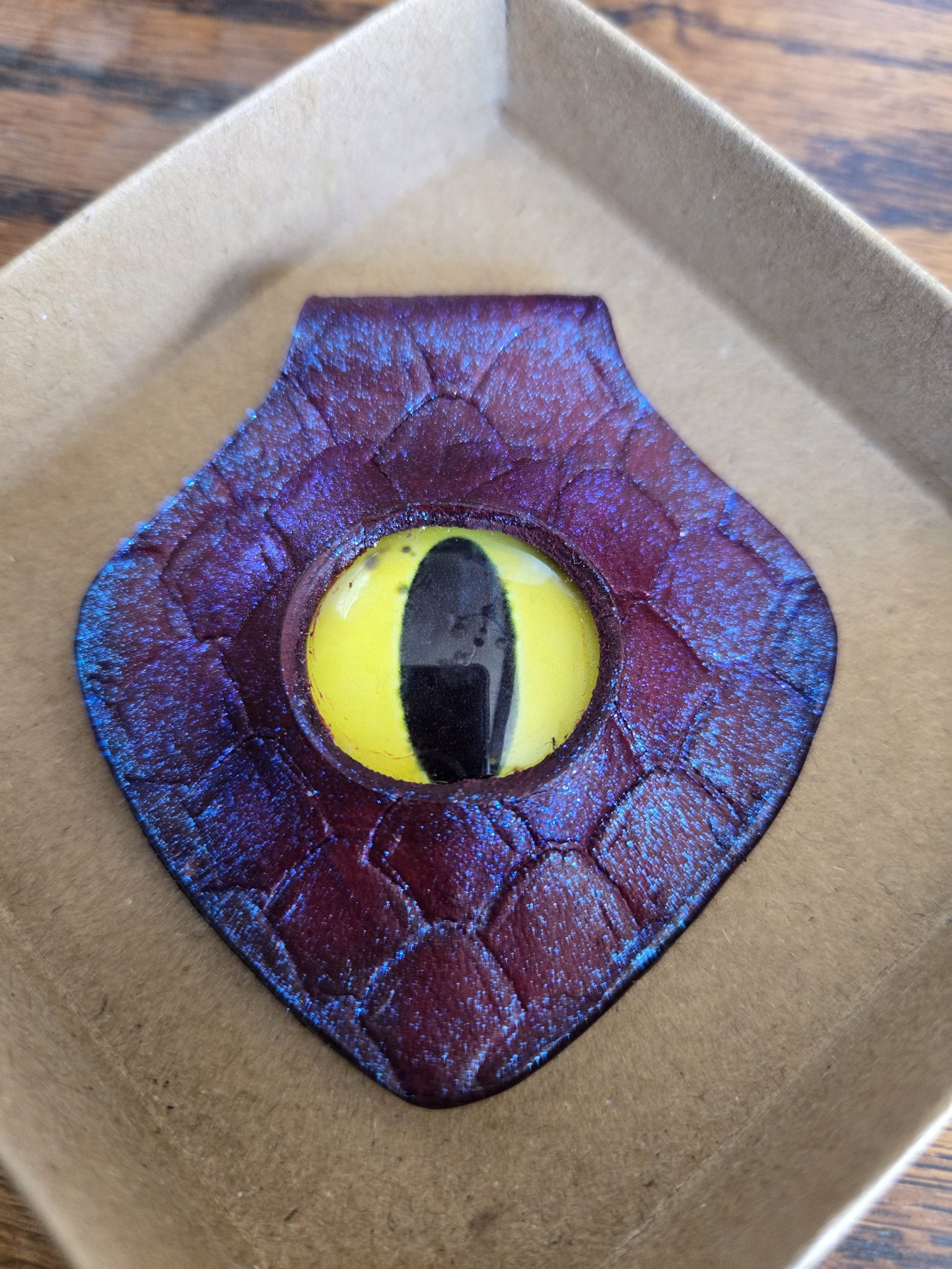 Stock 172 shield-shaped red leather pendant with dragon scale stamping, blue-purple color-change shimmer, and a yellow dragon eye cabochon, shown in a front close-up