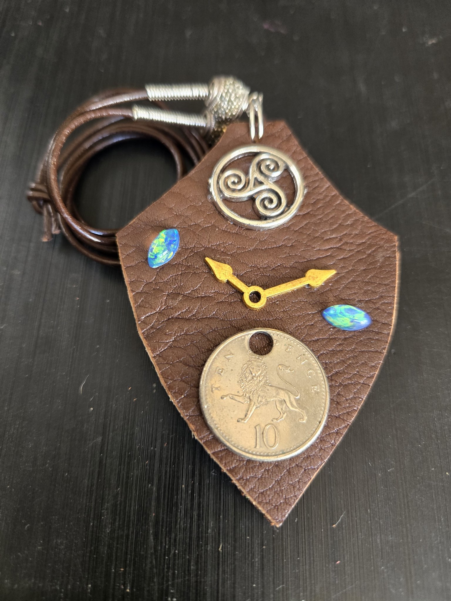Stock 178 shield-shaped brown buffalo leather pendant with a British 10 pence lion coin, silver-tone trinity medallion, gold-tone clock hands, and two blue-green holographic oval cabochons, shown with its cord