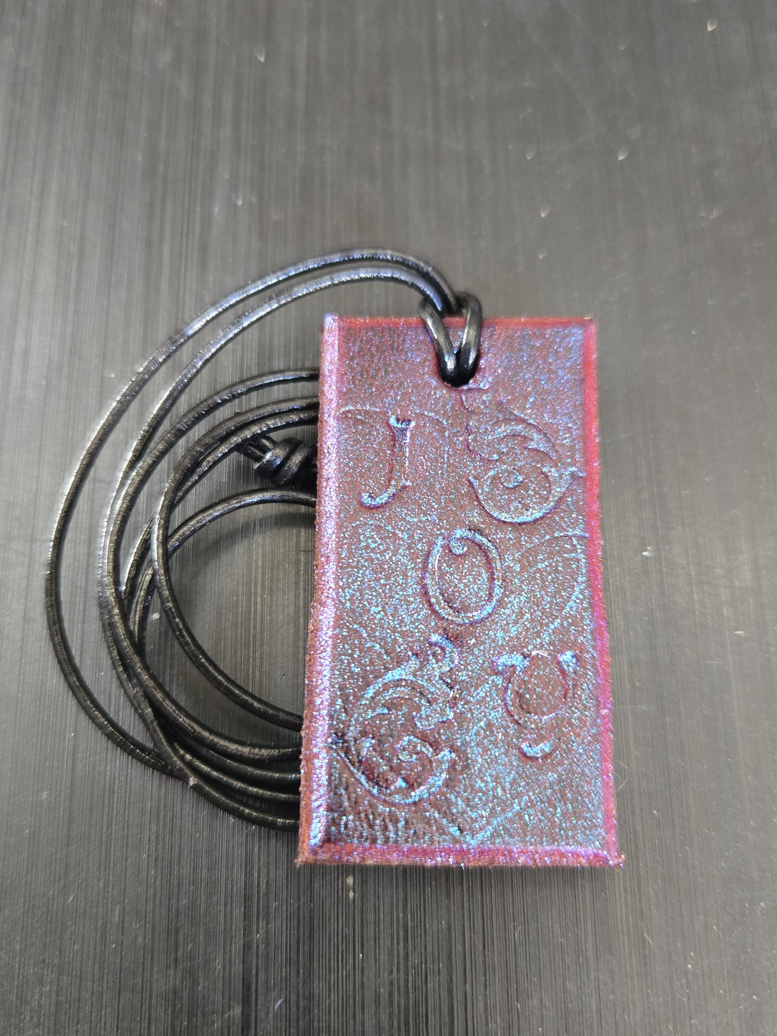 Stock 185 rectangular saddle leather pendant with blue-purple color-change shimmer and hand-stamped JOY lettering with floral scroll designs, shown in a front close-up