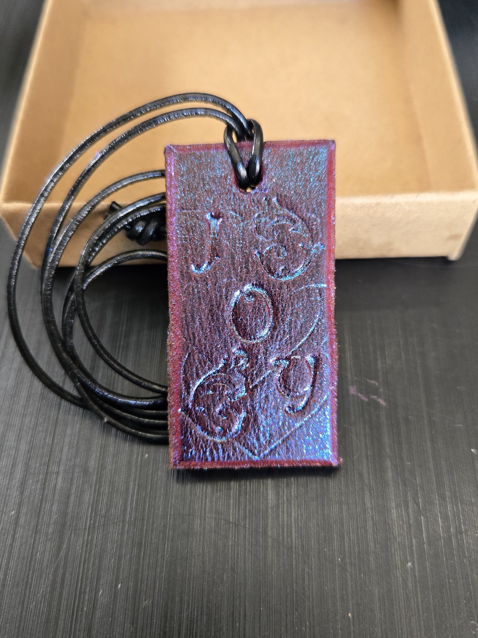 Front view of stock 185 JOY saddle leather pendant with its 28-inch black leather cord and a gift box in the background